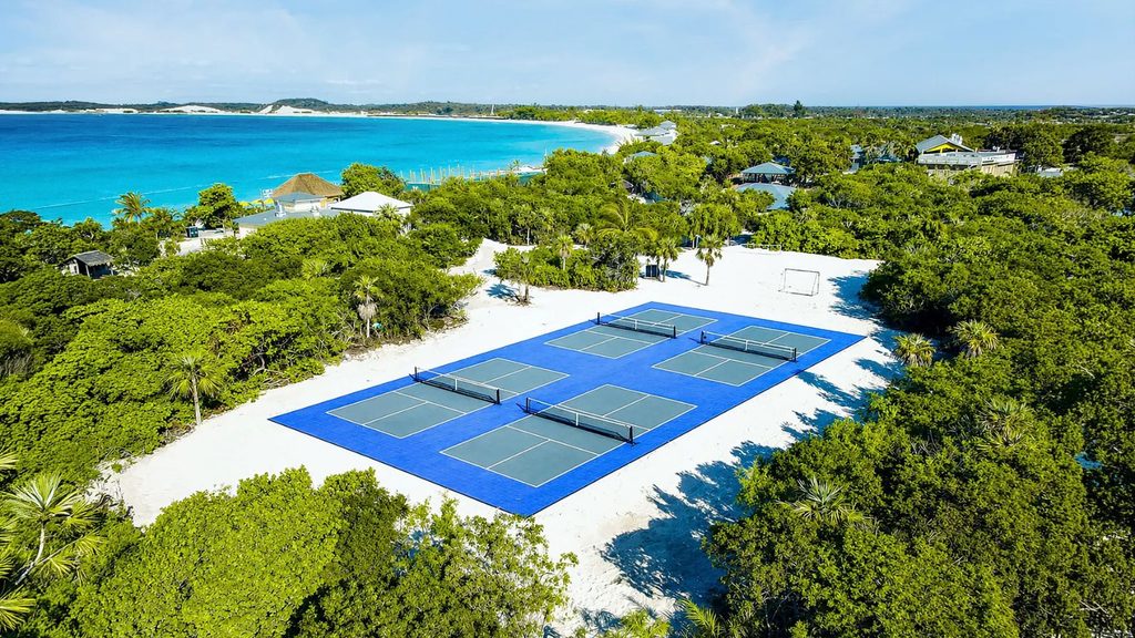 Aerial view of six outdoor pickleball courts on white sand near the ocean, surrounded by greenery and a few buildings on the picturesque Holland America private island.