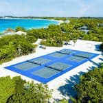 Aerial view of six outdoor pickleball courts on white sand near the ocean, surrounded by greenery and a few buildings on the picturesque Holland America private island.