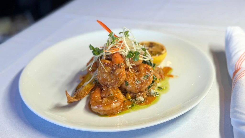 A white plate with cooked shrimp in sauce, garnished with microgreens and a grilled lemon half, on a table with a napkin beside it.