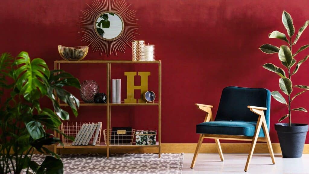 A modern room with a red wall, gold shelving, decorative items, a green armchair, indoor plants, a sunburst mirror, and a patterned rug.