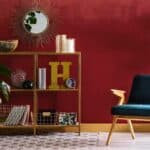 A modern room with a red wall, gold shelving, decorative items, a green armchair, indoor plants, a sunburst mirror, and a patterned rug.