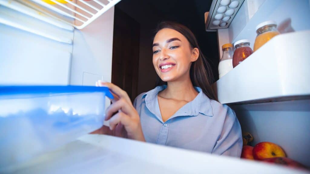 Woman smiling as she opens a container inside a refrigerator; food items and jars are visible on the fridge shelves.