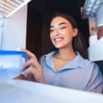 Woman smiling as she opens a container inside a refrigerator; food items and jars are visible on the fridge shelves.