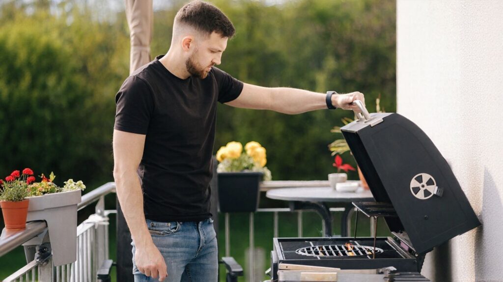 Man in a black t-shirt and jeans lifts the lid of a black grill on a balcony, with potted plants and outdoor furniture in the background.