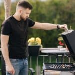 A man in a black t-shirt and jeans opens the lid of a grill on a patio with potted plants and outdoor furniture in the background.
