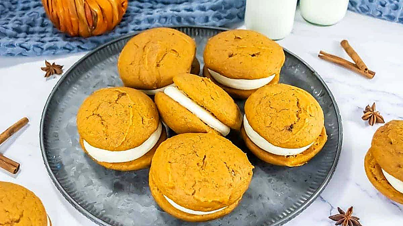 A plate with pumpkin whoopie pies filled with white cream, surrounded by cinnamon sticks, star anise, and a small pumpkin decoration.