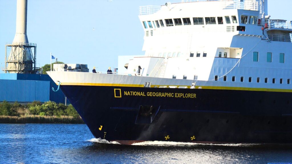 A large ship named &ldquo;National Geographic Explorer&rdquo; sails on the water near an industrial building and greenery under a clear sky.