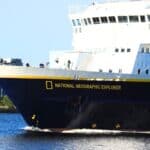 A large ship named “National Geographic Explorer” sails on the water near an industrial building and greenery under a clear sky.