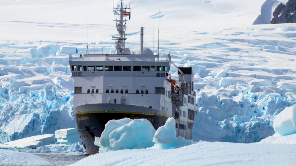 A large ship navigates through icy waters near a glacier, surrounded by icebergs and snow-covered terrain.