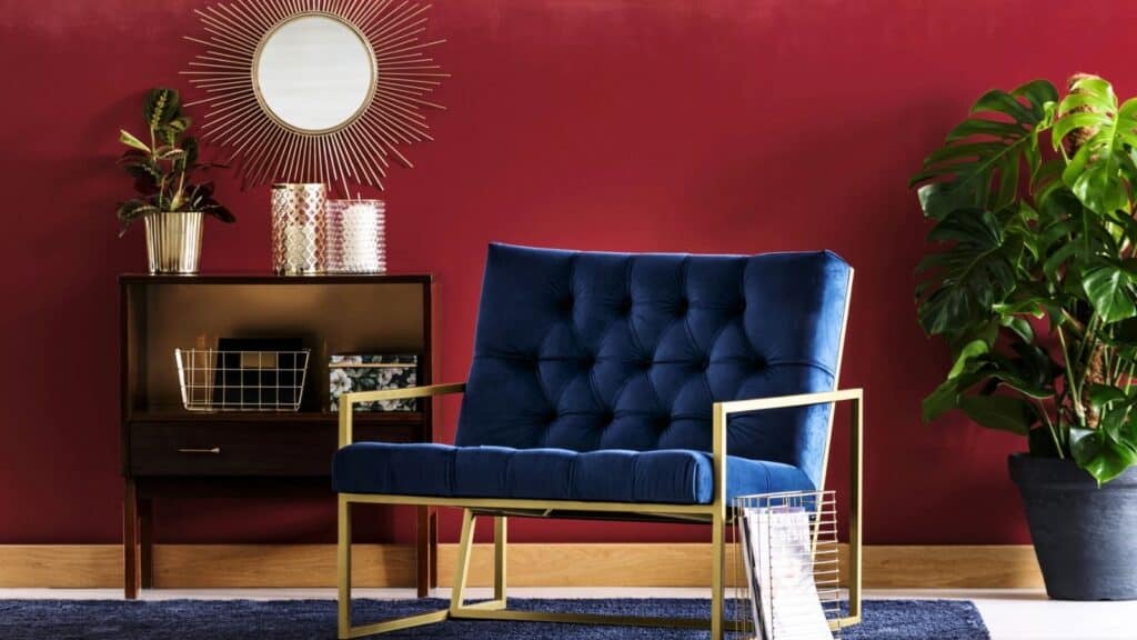 A blue velvet chair with gold frame sits by a wooden cabinet, a sunburst mirror, plants, and decor against a red wall with wood trim.