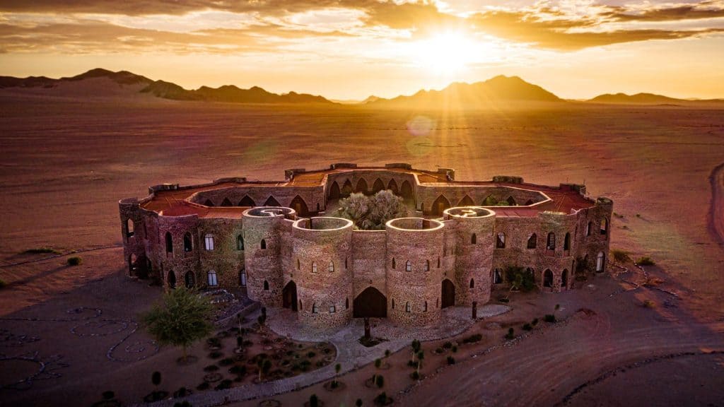 A large circular stone building sits in a desert landscape at sunrise, with mountains visible in the background.