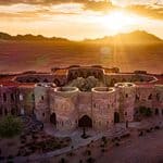 A large circular stone building sits in a desert landscape at sunrise, with mountains visible in the background.
