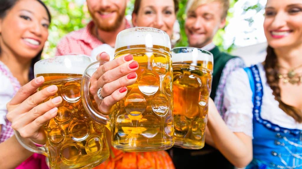 Five people in traditional Bavarian clothing hold large beer mugs, toasting together outdoors at Oktoberfest.