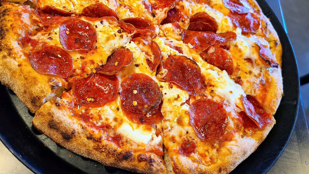 A close-up, top-down view of a round pepperoni pizza with melted cheese and red pepper flakes, served on a black tray—perfect for celebrating National Pepperoni Pizza Day.