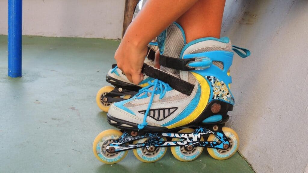 Person fastening the straps on a blue and gray inline skate while sitting on a hard, green surface, preparing to enjoy classic roller rink traditions.