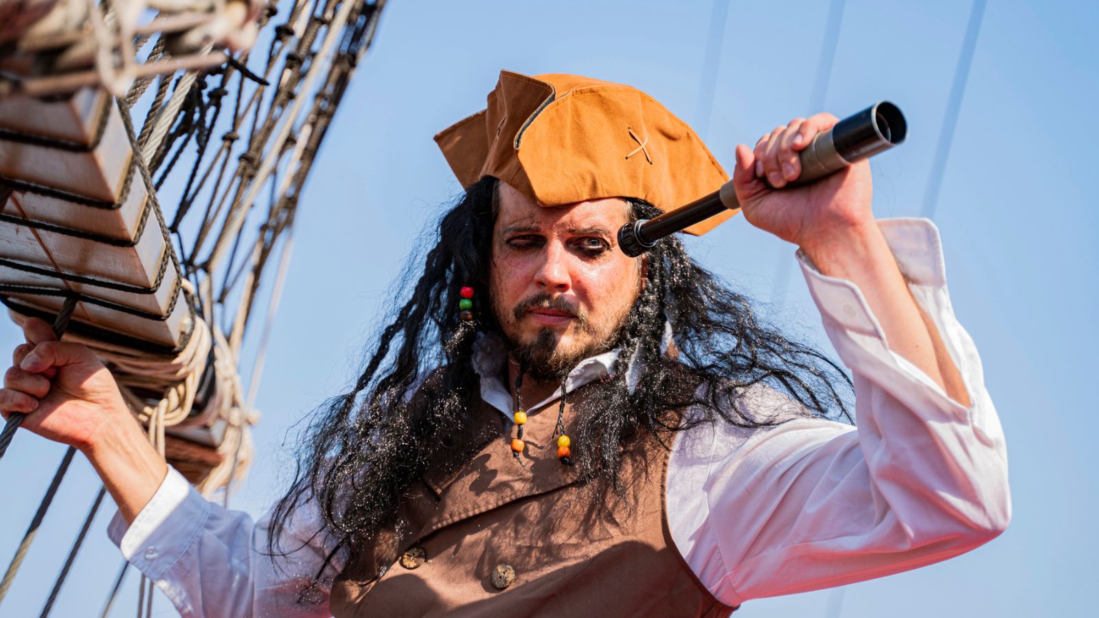 Person dressed as a pirate with a brown hat and vest holds a spyglass and stands on a ship, looking ahead&mdash;perfect for celebrating Talk Like a Pirate Day. Blue sky and ship ropes are visible in the background.