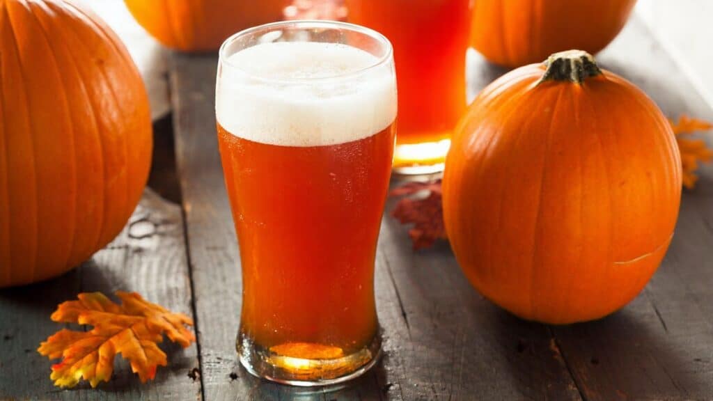 A pint glass of amber beer with foam sits on a wooden table surrounded by whole pumpkins and autumn leaves.