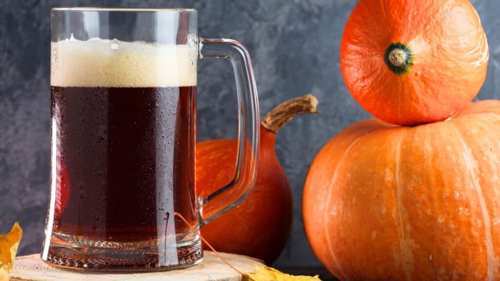 A glass mug of dark beer with a foamy head sits beside three orange pumpkins on a wooden surface.