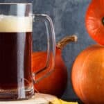 A glass mug of dark beer with a foamy head sits beside three orange pumpkins on a wooden surface.