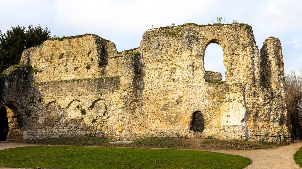 Stone ruins of Reading Abbey with arched windows and doorways, weathered walls, and patches of grass in the foreground under a partly cloudy sky.