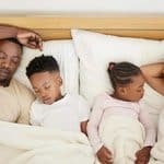 A family of four, two adults and two children, sleep side by side in bed under white blankets.