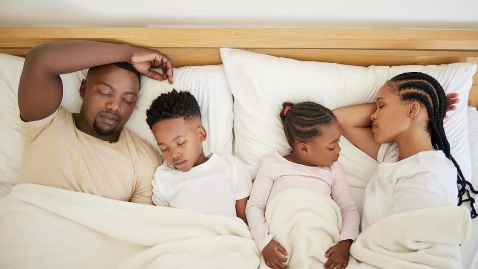 A family of four, two adults and two children, sleep side by side in bed under white blankets.