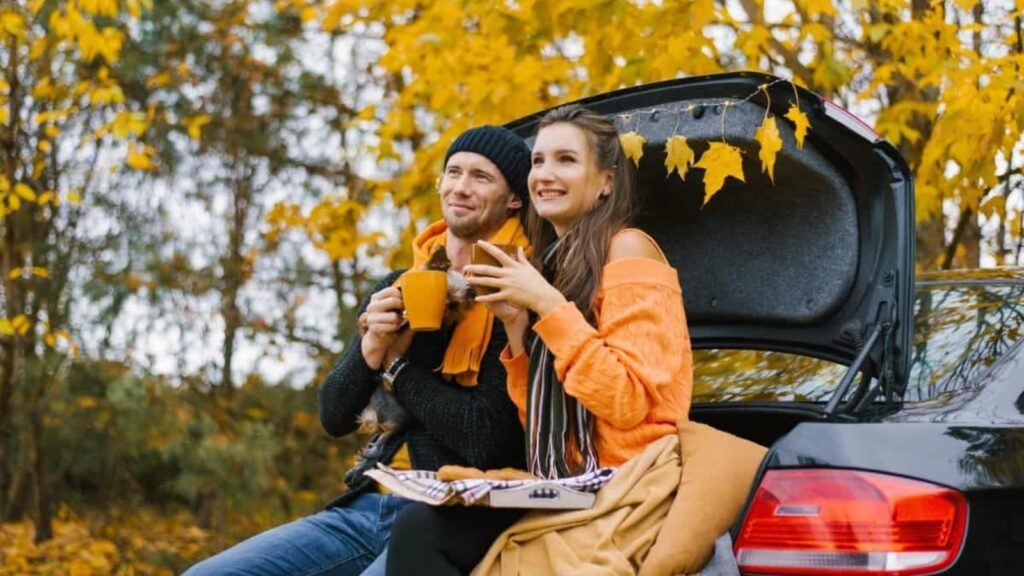 A man and woman sit on the open trunk of a car in an autumn forest, holding mugs and smiling, with yellow leaves in the background.