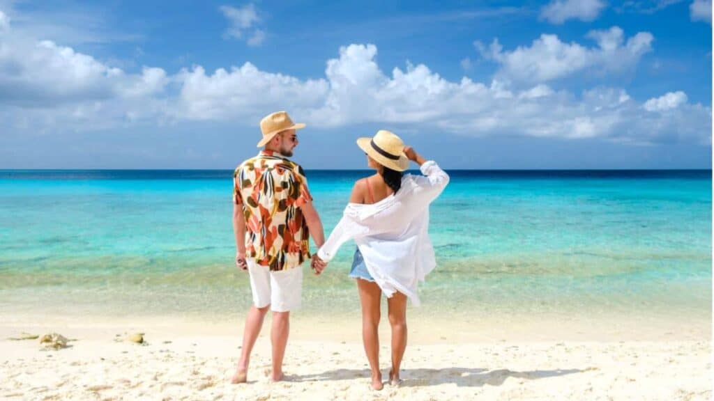 A couple stands on a sandy beach, holding hands and facing the turquoise ocean under a blue sky with scattered clouds, capturing the tranquil beauty often found at Caribbean resorts after Labor Day.