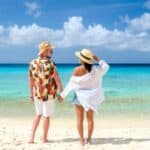 A couple stands on a sandy beach, holding hands and facing the turquoise ocean under a blue sky with scattered clouds, capturing the tranquil beauty often found at Caribbean resorts after Labor Day.