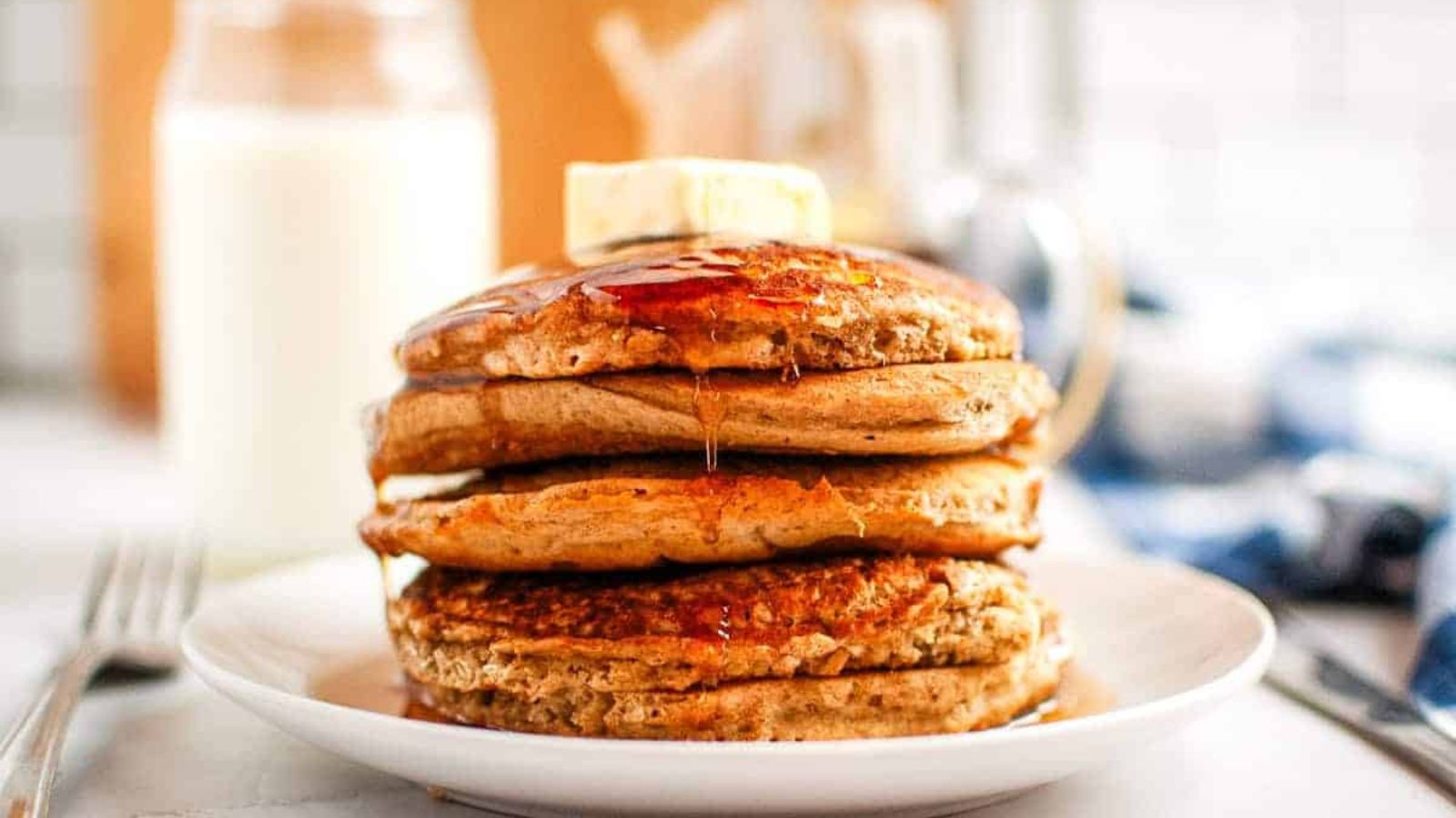 A stack of pancakes topped with butter and syrup on a white plate, with a glass bottle of milk in the background.
