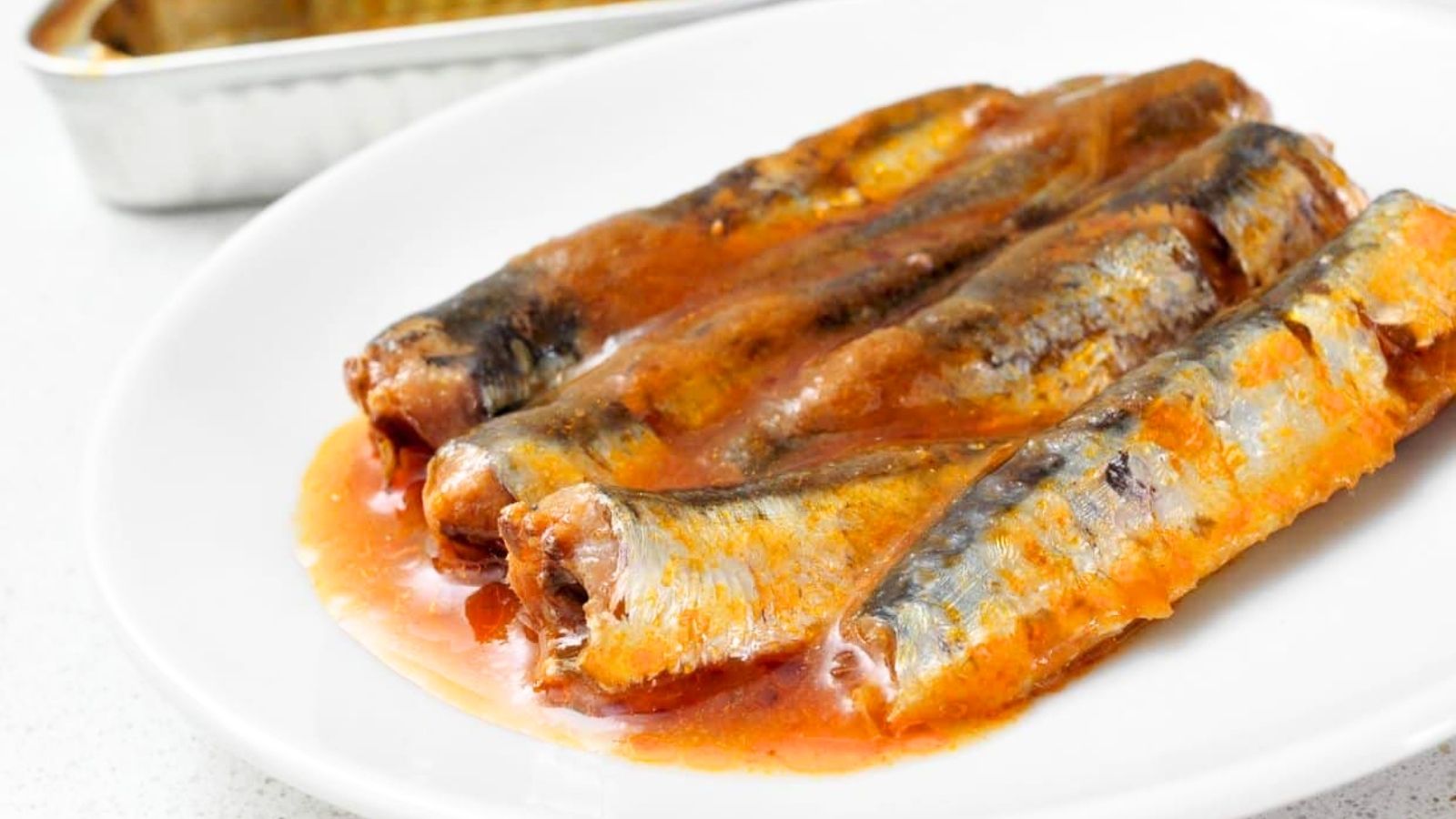 Four sardines in tomato sauce are arranged on a white plate, with some sauce pooling around them. A metal tray is partially visible in the background.