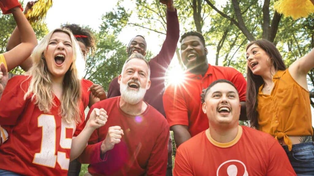 A group of people outdoors wearing red and yellow shirts cheer enthusiastically, with sunlight shining through trees in the background.