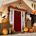 A house with a red door is decorated for Halloween with carved pumpkins, gourds, hay bales, black cloth, crows, and fall leaves on the porch.