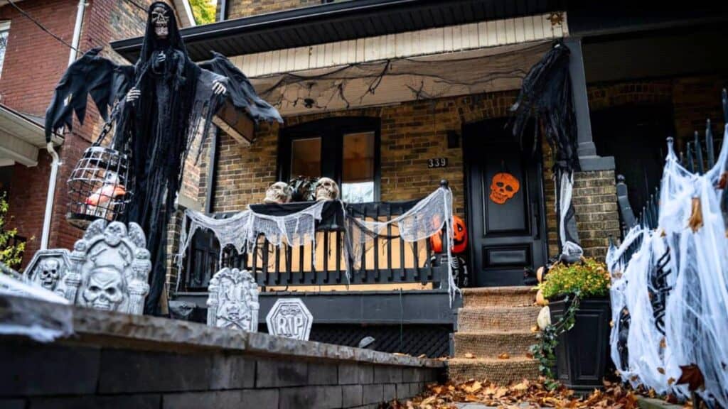 A house porch decorated for Halloween with skulls, fake cobwebs, tombstones, hanging ghouls, and orange pumpkin decorations.