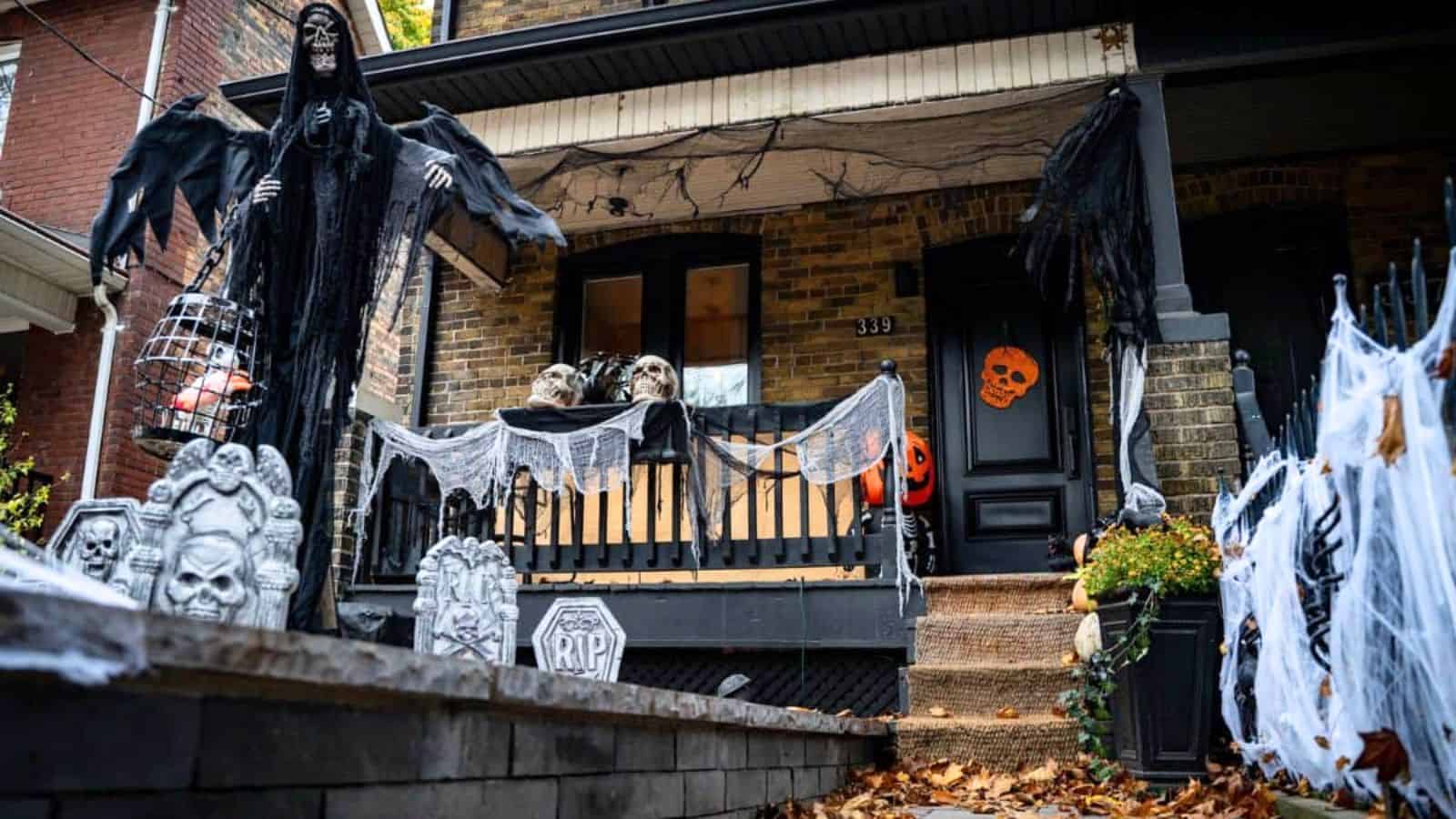 A house porch decorated for Halloween with skulls, fake cobwebs, tombstones, hanging ghouls, and orange pumpkin decorations.
