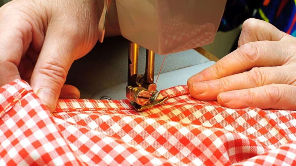 Hands guiding red and white checkered fabric through a sewing machine, stitching along the edge.