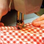 Hands guiding red and white checkered fabric through a sewing machine, stitching along the edge.