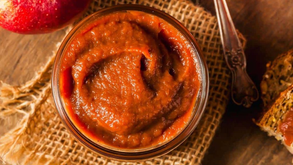 A glass jar filled with thick, dark brown apple butter sits on burlap beside an apple and a slice of bread.