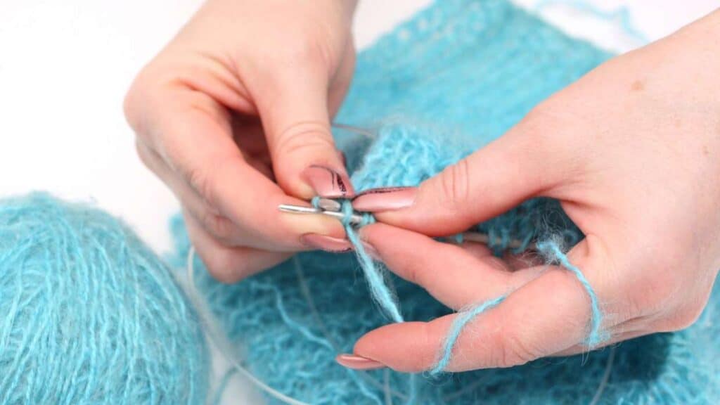 Close-up of hands knitting with light blue yarn and metal needles; a partially completed knitted fabric and a ball of yarn are visible.