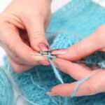 Close-up of hands knitting with blue yarn and metal needles, capturing the creativity often found in craft studios. A ball of blue yarn and knitted fabric are visible in the background.