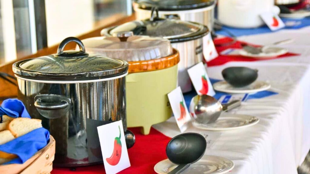 Four slow cookers line a buffet table with ladles, plates, and chili pepper cards indicating spice levels; a basket of bread is on the side.