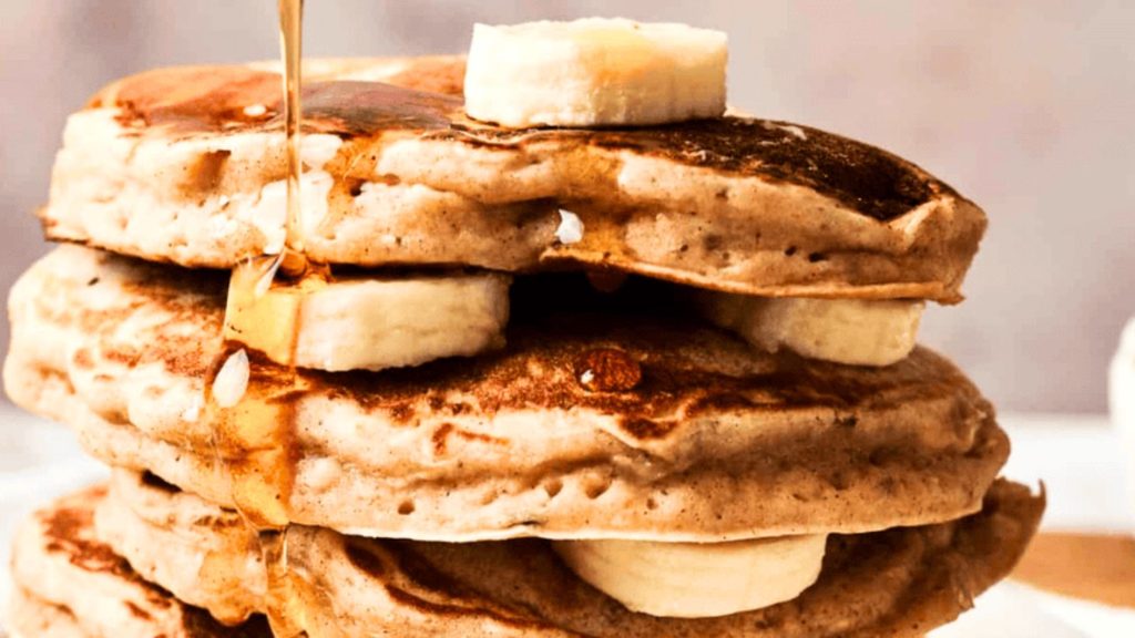 A stack of pancakes with banana slices between layers, topped with syrup being poured over them.