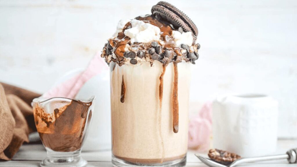 A glass of milkshake topped with whipped cream, chocolate syrup, chocolate chips, and a cookie, with a spoon and a small pitcher of chocolate sauce beside it.