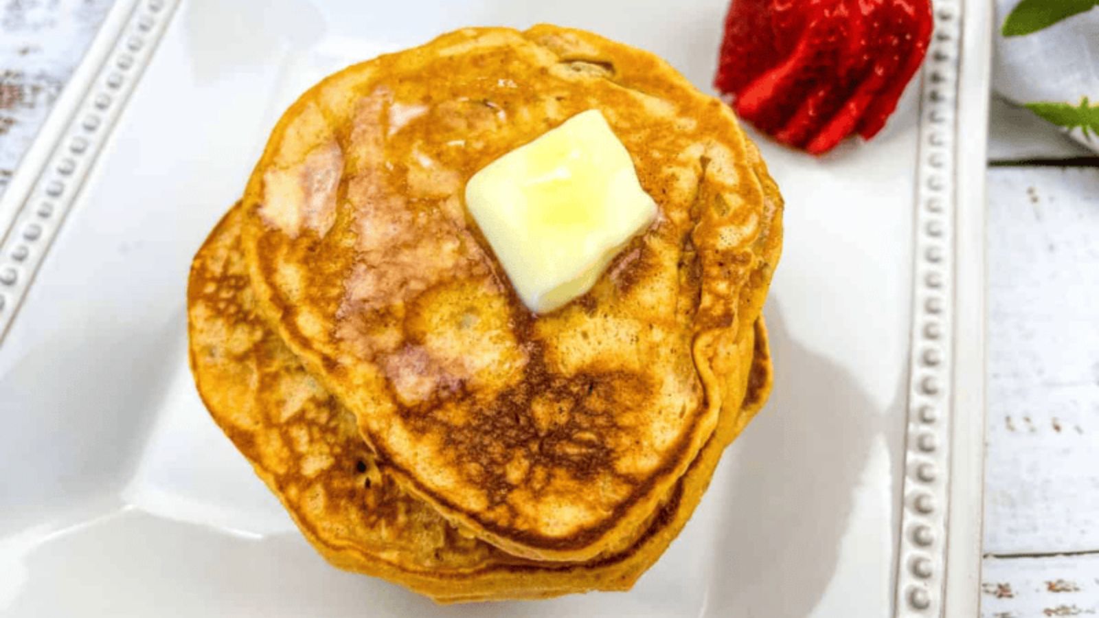 A stack of pancakes with a pat of melting butter on top, served on a white plate with a sliced strawberry on the side&mdash;perfect for celebrating National Pancake Day.