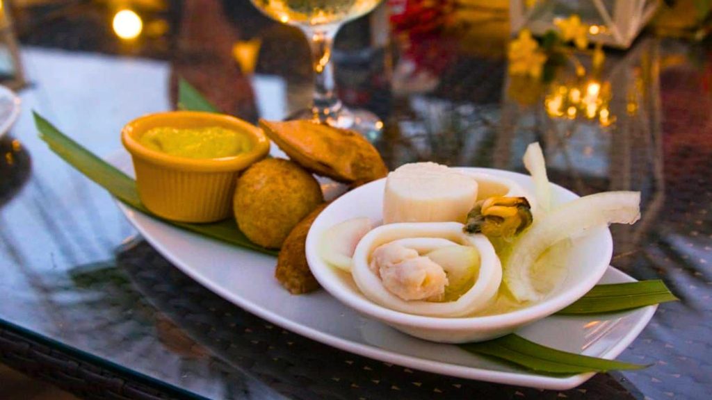 A white plate with seafood, onions, a mussel, fried snacks, and a small dish of yellow sauce on a glass table.