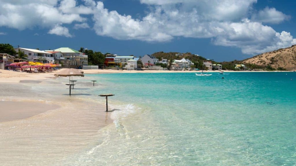 Sandy beach with clear turquoise water, beach umbrellas, and tables; small houses and hills in the background under a partly cloudy sky make this the perfect setting for a relaxing fall beach trip.