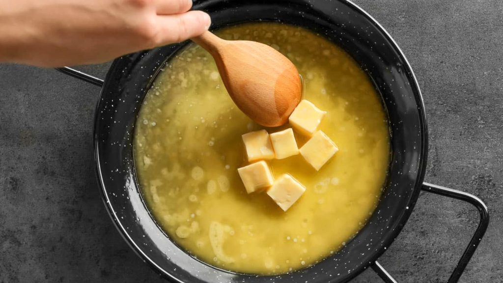 A hand holding a wooden spoon stirs cubed butter melting into a fragrant brown butter in a yellow liquid in a black pan on a gray surface.