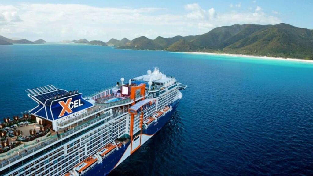 A large cruise ship labeled “Xcel” sails near a tropical coastline with clear blue water and green mountains under a partly cloudy sky.