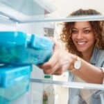A woman smiles while reaching for a blue food container inside a refrigerator, surrounded by other similar containers.