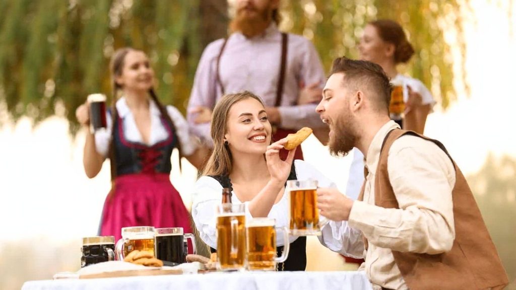 Two people sit at a table outdoors sharing food and beer, while three others in traditional attire stand in the background holding mugs.